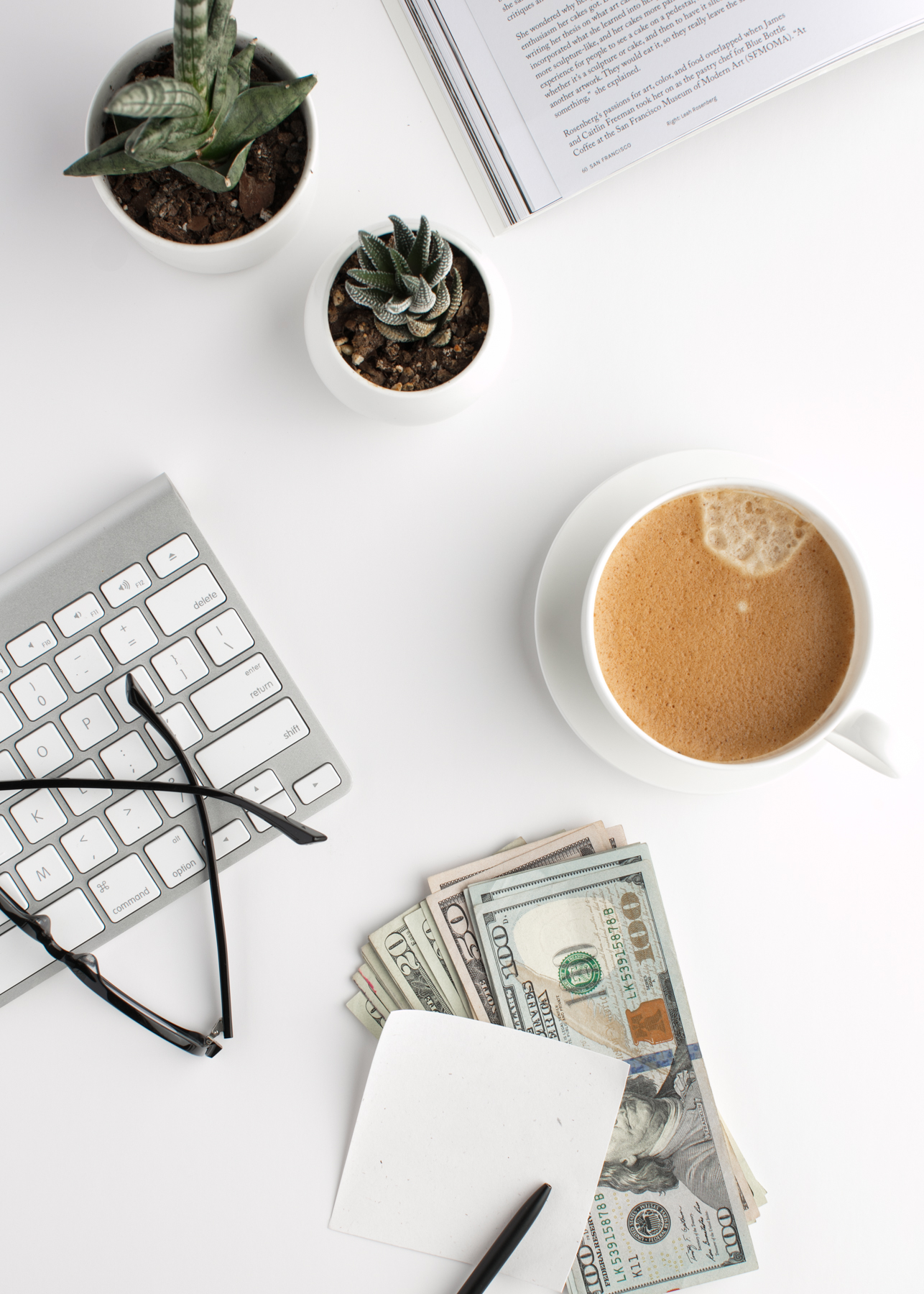 flat lay of a keyboard, coffee, reading glasses and a stack on 100 dollar bills.